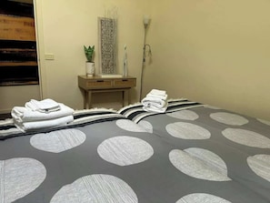 Een keurig opgemaakt bed met een grijs-wit stippenbedover, een nachtkastje met een plant, en een wandlamp.