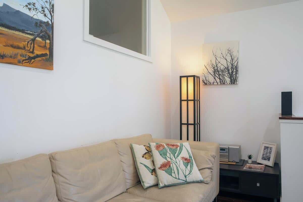 Ein Wohnzimmer mit einem beigen Sofa, zwei Zierkissen, einer Stehlampe, einem Gemälde und einem kleinen Tisch mit Radio und Bilderrahmen.