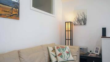 Ein Wohnzimmer mit einem beigen Sofa, zwei Zierkissen, einer Stehlampe, einem Gemälde und einem kleinen Tisch mit Radio und Bilderrahmen.