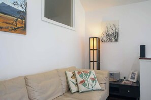 Un salon avec un canapé beige, deux coussins décoratifs, une lampe sur pied, un tableau et une petite table sur laquelle se trouvent une radio et un cadre photo.