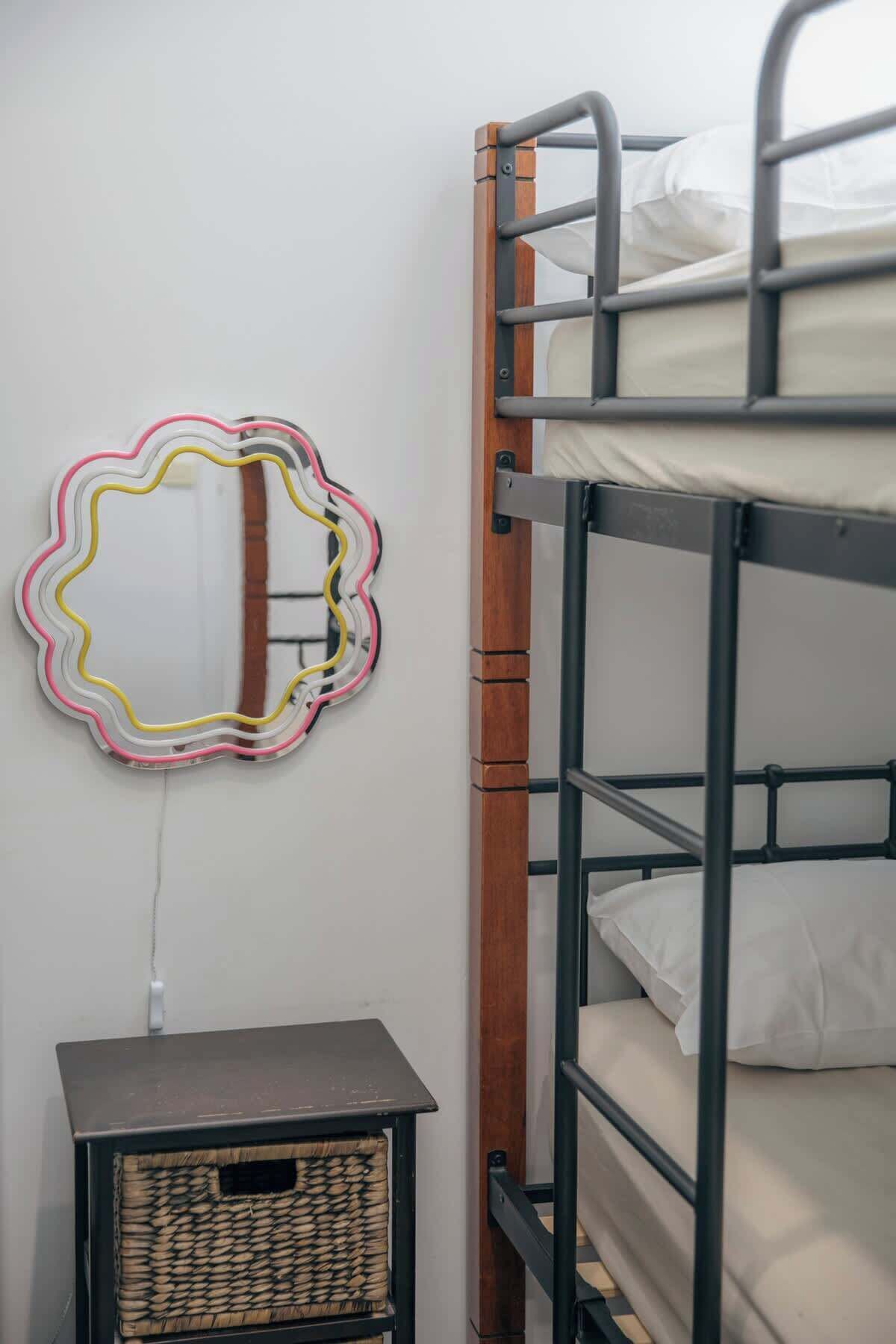 Ein Stockbett mit Holzrahmen und weißer Bettwäsche, ein kleiner Nachttisch mit einem gewebten Korb und ein an der Wand befestigter Spiegel mit farbigem Rahmen.