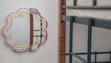 Ein Stockbett mit Holzrahmen und weißer Bettwäsche, ein kleiner Nachttisch mit einem gewebten Korb und ein an der Wand befestigter Spiegel mit farbigem Rahmen.
