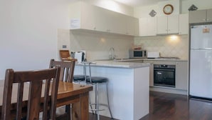 A kitchen with white cabinets, a microwave, a refrigerator, and a dining area with a wooden table and chairs. - Halls Gap Escape Townhouse 6 (Halls Gap)
