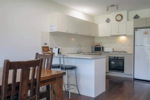 A kitchen with white cabinets, a microwave, a refrigerator, and a dining area with a wooden table and chairs. - Halls Gap Escape Townhouse 6 (Halls Gap)