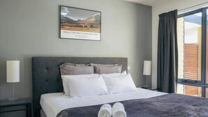 Ein Hotelzimmer mit einem großen Bett, zwei Nachttischen, einem fensterbelaubten Fenster und einem gerahmten Bild an der Wand.