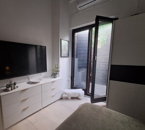 A modern bedroom with a flat-screen TV on a white dresser, a small stool, and a sliding glass door leading to an outdoor area. - The Village Retreat
Live like a local! (Malonas)