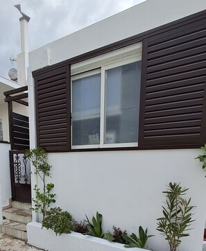A building with a white exterior, a window with dark shutters, and a small garden with various plants. - The Village Retreat
Live like a local! (Malonas)