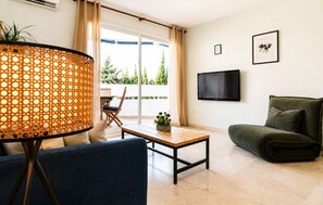 A living room with a sofa, a table, a lamp, a TV, and a balcony with a view of trees. - Coastal Living Near Estepona Marina (Estepona)