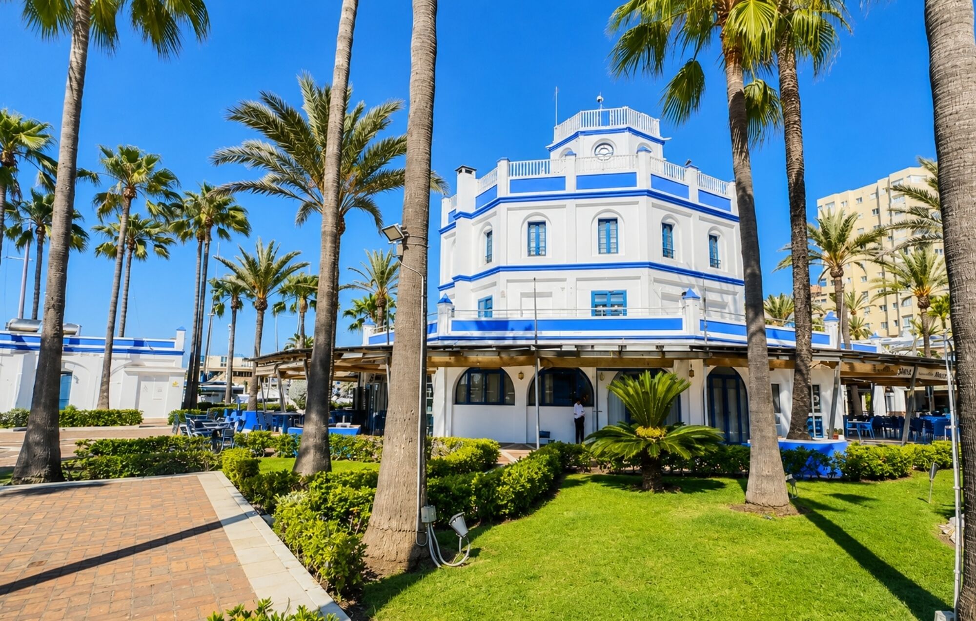 A white building with blue trim and a clock tower, surrounded by palm trees and a paved walkway.