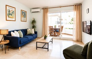Ein Wohnzimmer mit blauem Sofa, Couchtisch, grünem Sessel, Fernseher und einem Balkon mit Tisch und Stühlen.