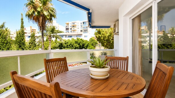 A round wooden table with four chairs, a potted plant, and a glass balustrade overlooking a residential area with palm trees and buildings. - Coastal Living Near Estepona Marina (Estepona)