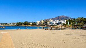 A beach with a wooden walkway leading to the water, a pool area, and buildings in the background. - Coastal Living Near Estepona Marina (Estepona)
