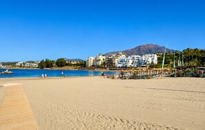 A beach with a wooden walkway leading to the water, a pool area, and buildings in the background. - Coastal Living Near Estepona Marina (Estepona)