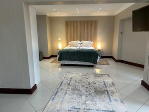 A bedroom with a bed, two bedside lamps, a large rug, and a wooden headboard. - Menlyn Stay (Pretoria)