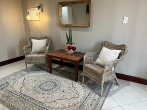 A room with two wicker chairs, a wooden coffee table, a patterned rug, a wall-mounted light fixture, and a mirror.