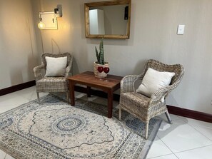 A room with two wicker chairs, a wooden coffee table, a patterned rug, a potted plant, a wall-mounted light fixture, and a mirror.