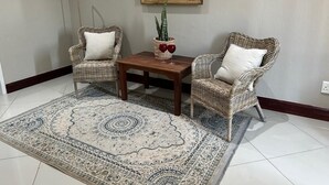A room with two wicker chairs, a small wooden table, a patterned rug, a potted plant, and a wall-mounted mirror. - Menlyn Stay (Pretoria)