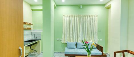 A room with a sofa, a small table with a vase, a wooden chair, a kitchen area with a sink, and a window with curtains.