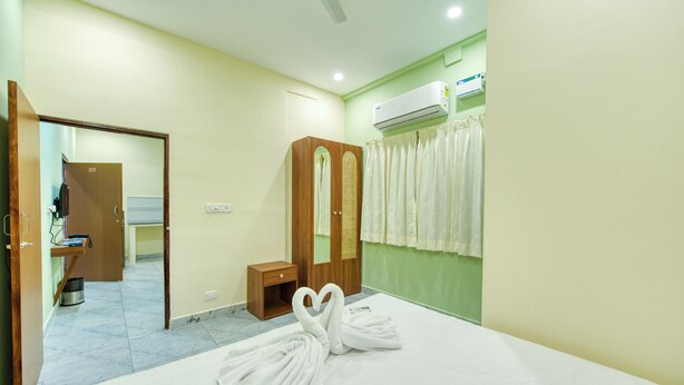 A hotel room with a bed, a wooden wardrobe, a small bedside table, a mirror, an air conditioning unit, and a bathroom with a shower curtain.