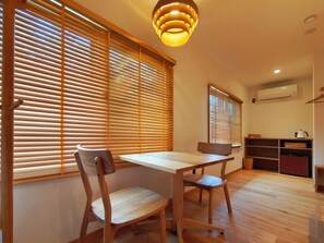 Un espace repas avec une table et des chaises en bois, de grandes fenêtres équipées de stores, un luminaire suspendu et un coin cuisine avec des étagères et un climatiseur.