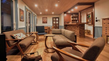 Una sala de estar acogedora con sofá, sillas y una pequeña mesa. Hay una zona de cocina con barra y estantes llenos de botellas.