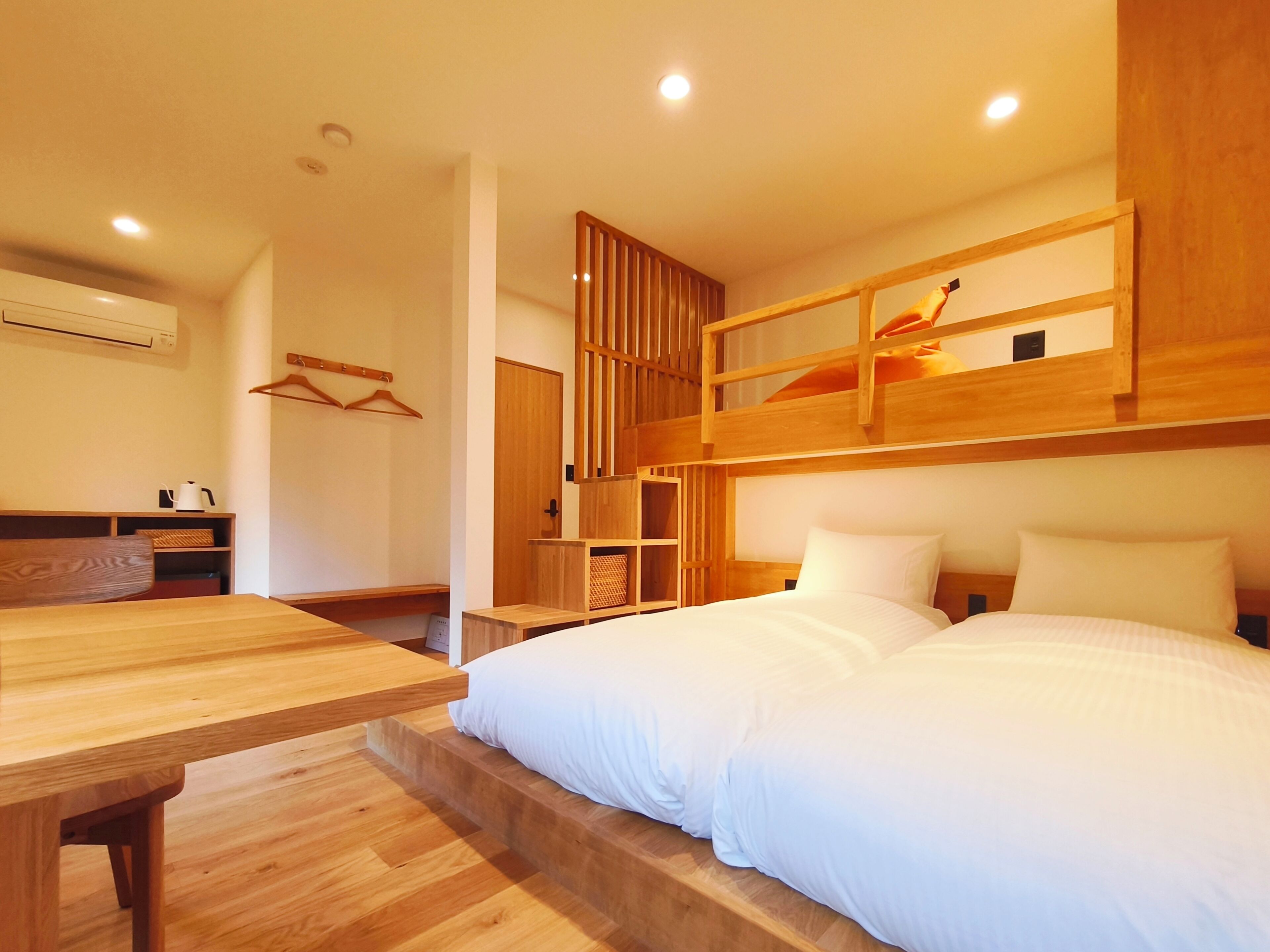 A room with a bunk bed, a wooden desk, a hanging coat rack, and a small kitchenette area.