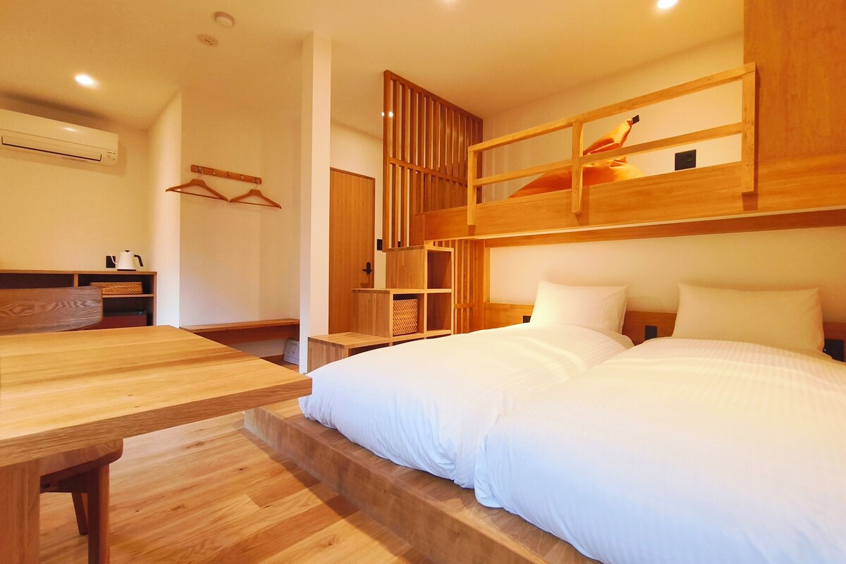 Una habitación con una cama litera, un escritorio de madera, un perchero y una pequeña zona de kitchenette.