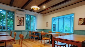 A dining area with wooden tables and chairs, large windows with views of greenery, and artwork on the walls.
