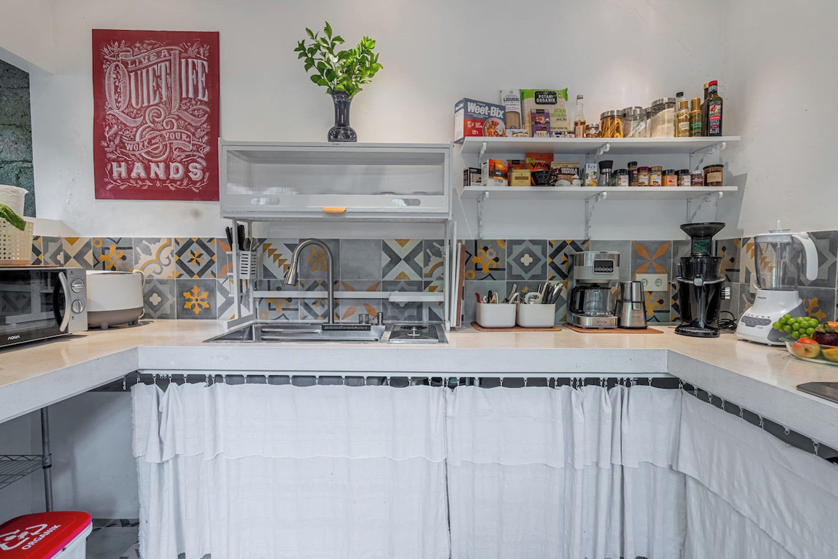 A kitchen with a microwave, sink, and various appliances. There is a plant on the counter and a poster on the wall.