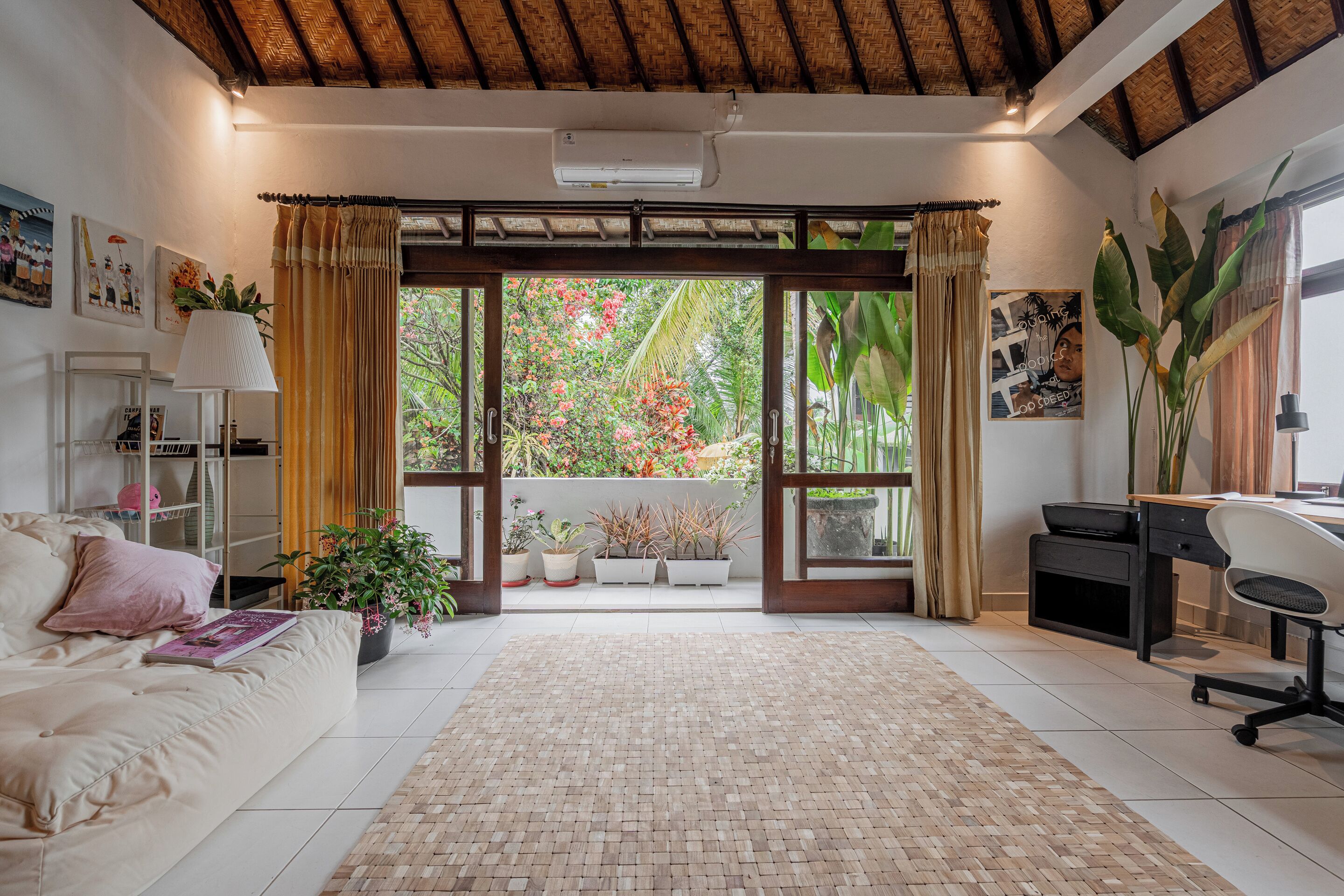 Una habitación con sofá, escritorio, computadora y vista a un jardín a través de un ventanal.