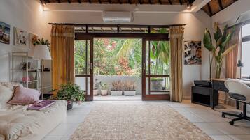Una habitación con sofá, escritorio, computadora y vista a un jardín a través de un ventanal.