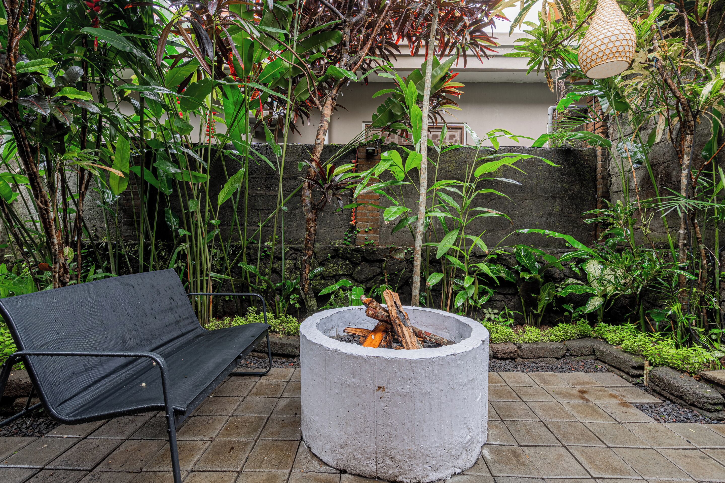 A concrete fire pit surrounded by lush greenery and a black metal bench.