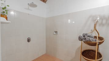 Un baño con azulejos blancos, una ducha con cabezal cromado, un perchero con toallas enrolladas y una planta en maceta a la izquierda.