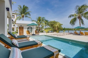 Un espace aménagé au bord de la piscine avec des chaises longues, un petit bâtiment sur lequel est apposée une enseigne, et des palmiers.