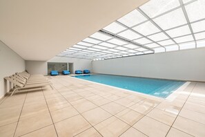 Una piscina interior con techo de tragaluces, suelo de baldosas y sillones azules.
