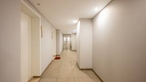 Un long couloir étroit, typique d’un hôtel, avec des portes sur le côté gauche, une moquette rouge et un sol carrelé.