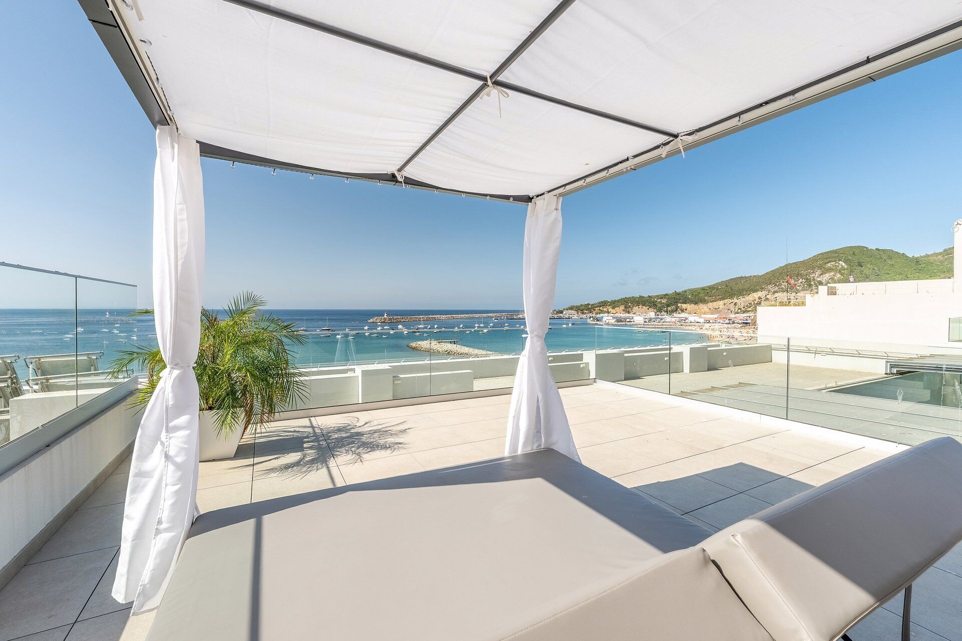 A balcony with a view of the sea, white curtains, and a glass table.