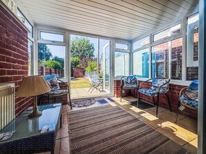 Quarto iluminado pelo sol com uma porta de vidro que dá para um jardim, mobiliado com cadeiras e uma mesa.