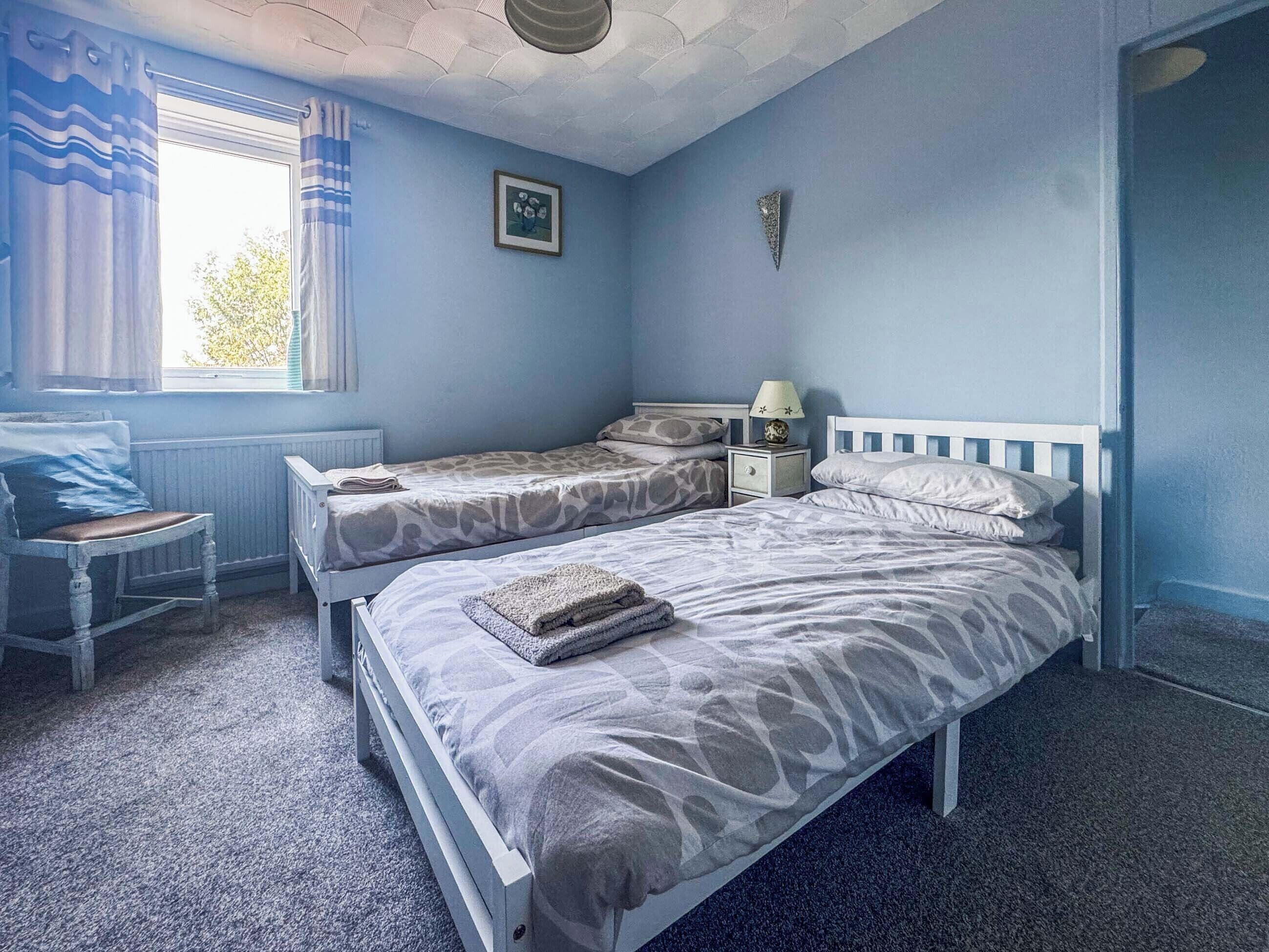 A bedroom with two beds, a window with curtains, a bedside table with a lamp, and a radiator.