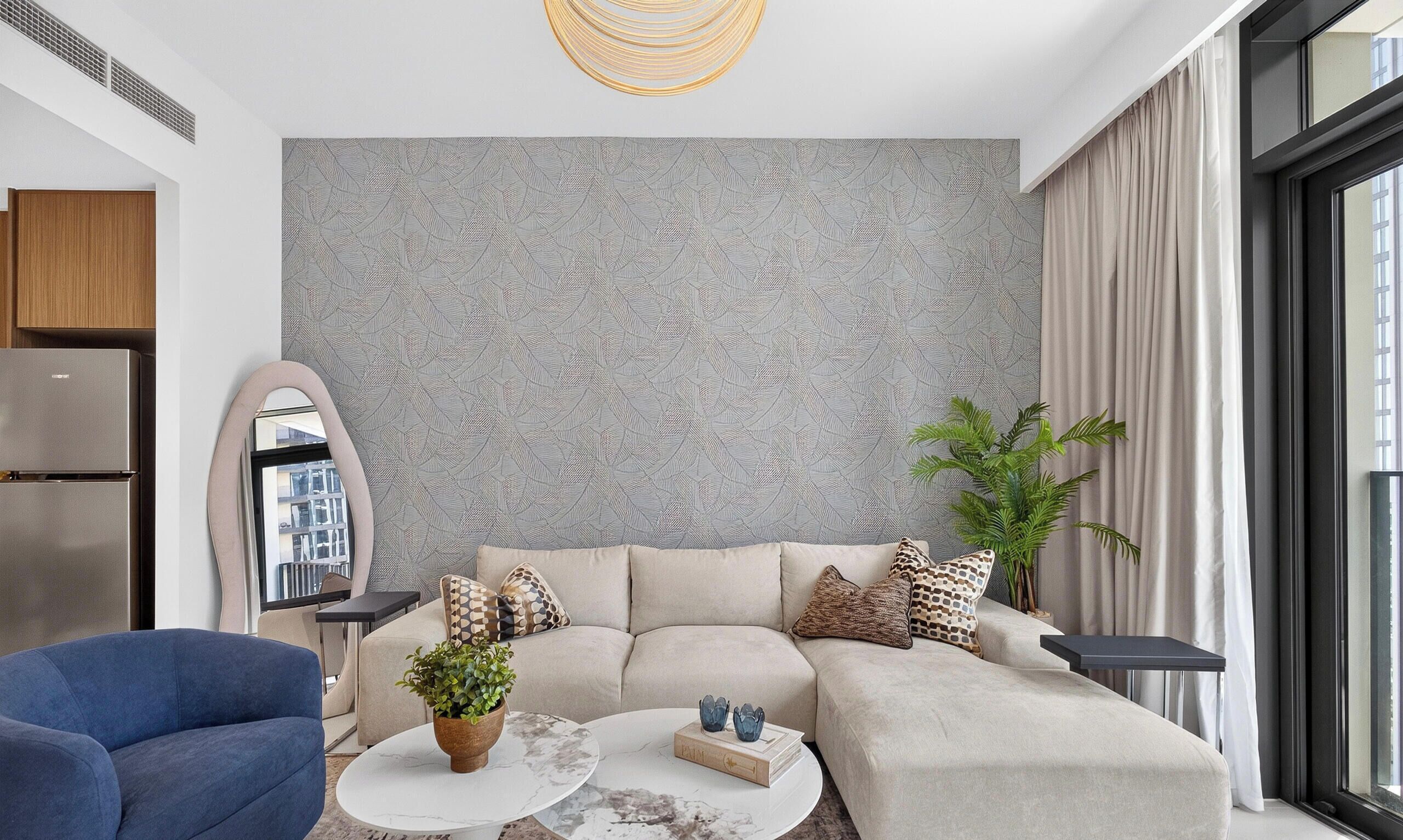 Un salon moderne avec un canapé d’angle, une table basse, un fauteuil bleu, une plante en pot et une grande fenêtre.