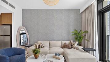 Un salon moderne avec un canapé d’angle, une table basse, un fauteuil bleu, une plante en pot et une grande fenêtre.
