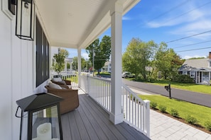Ein überdachter Balkon mit einem Korbsessel und einem Tisch, mit Blick auf eine Straße mit Häusern und Grünflächen.