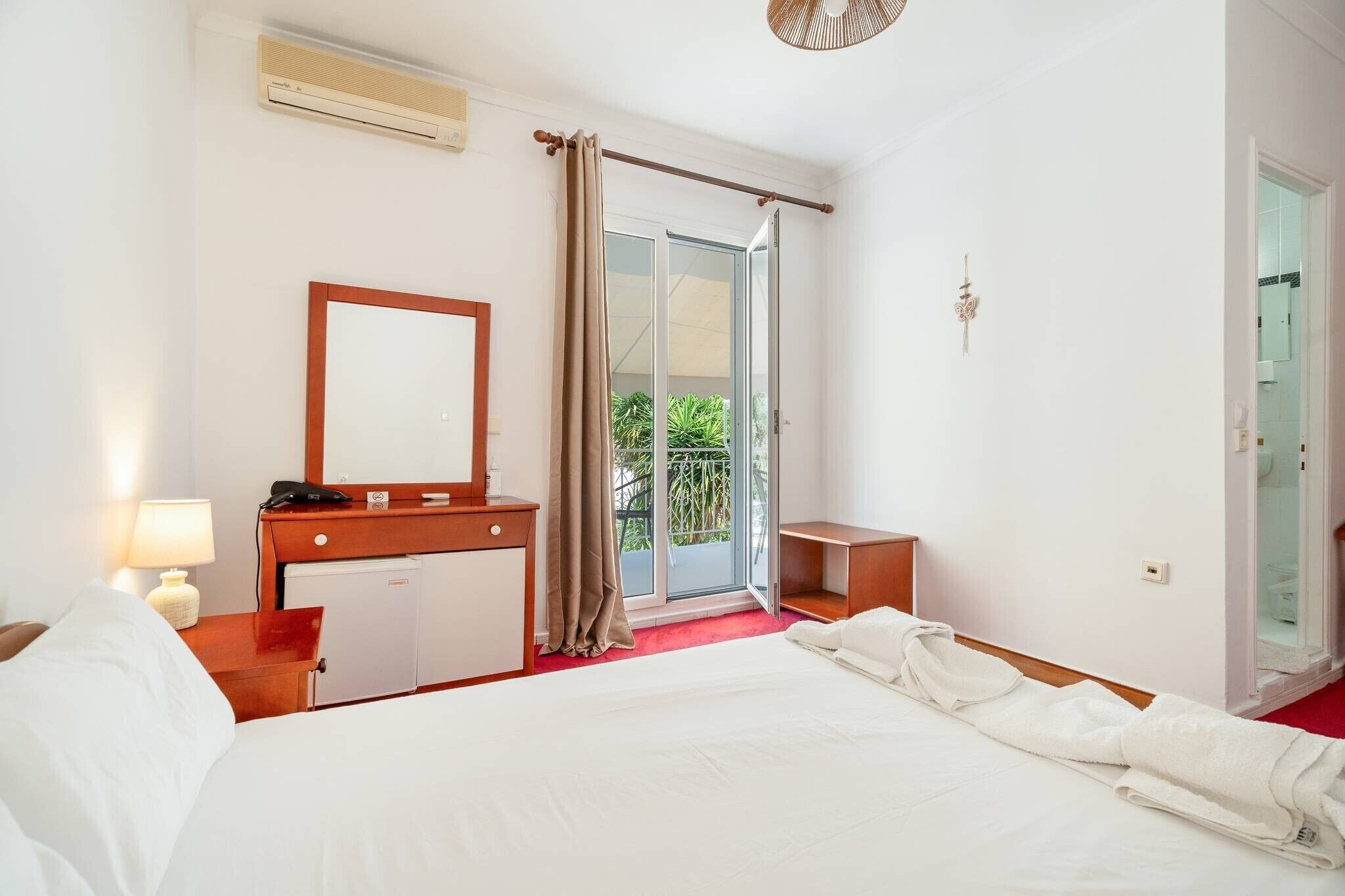 Une chambre d’hôtel comprenant un lit, un bureau avec un miroir, un petit réfrigérateur, une table de chevet avec une lampe et une porte-fenêtre coulissante donnant sur le balcon.