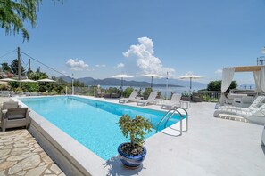 Piscina com espreguiçadeiras e guarda-sóis, rodeada por um deck de pedra e vista para montanhas.