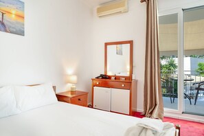 Quarto de hotel com cama, escrivaninha com luminária, minibar e uma janela grande com cortinas.