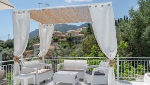 A balcony with white wicker furniture, a canopy, and a view of houses and mountains. - Room 'Ground Floor' Shared Pool and Wi-Fi (Nikiana)