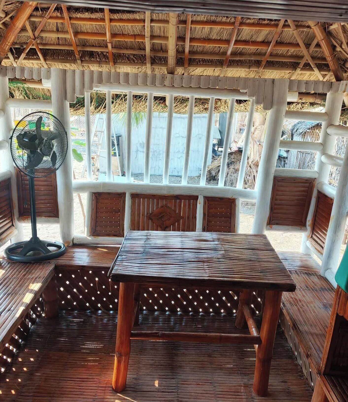 A wooden table and bench set under a thatched roof structure with a view of a beach.