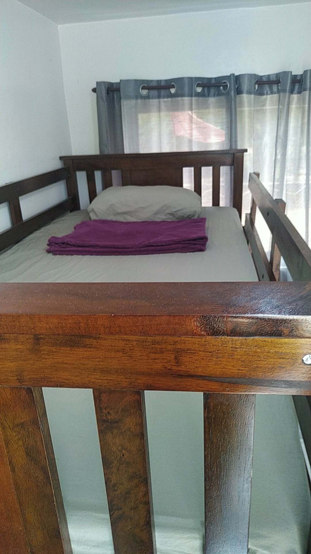 A single bed with a wooden headboard and a purple blanket, a window with curtains, and a white wall.