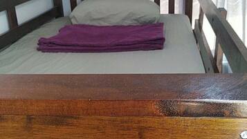 A single bed with a wooden headboard and a purple blanket, a window with curtains, and a white wall.
