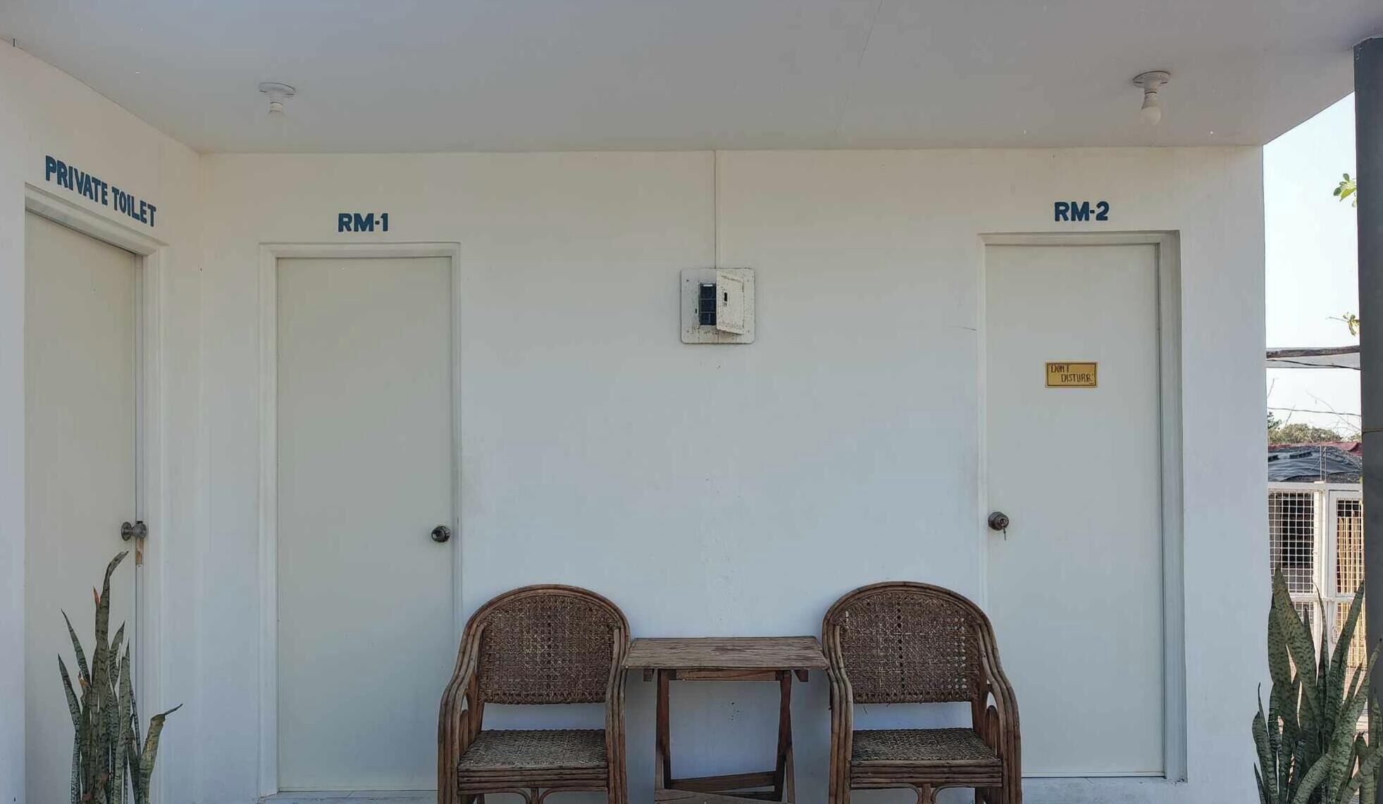 Un espace toilettes privatif comprenant deux portes portant les numéros RM-1 et RM-2, une petite table, deux chaises en osier et une plante en pot.